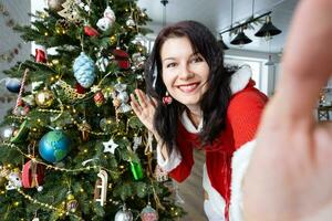 Selfie portrait of a woman in a red Santa dress in a home interior with a Christmas tree and New Year decor. Preparation for the holidays, party photo