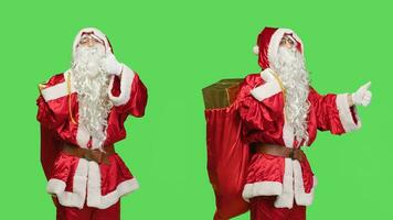 Father christmas shows thumbs up on camera, feeling positive giving like and agreement symbol over greenscreen backdrop. Santa claus in suit expressing approval with gifts bag. photo