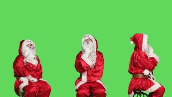 Santa on chair brainstorm gift ideas sitting over greenscreen backdrop, thinking about presents before christmas eve holiday celebration. Young man dressed as saint nick in studio. photo