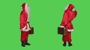 Father christmas carries suitcase in studio, wearing traditional festive costume and red hat. Saint nick character with briefcase posing onver greenscreen backdrop, hitchhiking. photo