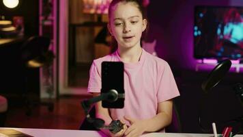 Kid films with smartphone attached to selfie stick in pink neon lit living room used as professional studio. Young media star captures footage with cellphone camera, discussing about fun day at school video
