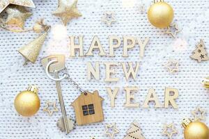 House key with keychain cottage on festive knitted background with stars, lights of garlands. Happy New Year wooden letters, greeting card. Purchase, construction, relocation, mortgage, insurance photo