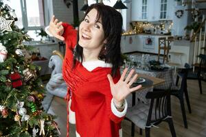 Selfie portrait of a woman in a red Santa dress in a home interior with a Christmas tree and New Year decor. Preparation for the holidays, party photo