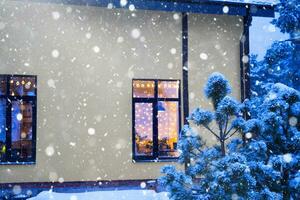 acogedora ventana festiva de la casa exterior con la cálida luz de las guirnaldas de luces de hadas en el interior: celebre la navidad y el año nuevo en un hogar cálido. árbol de navidad, bokeh, nieve en pinos y nevadas foto