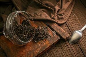 Dried leaf black tea is scattered from glass jars on a wooden vintage board with a spoon and a cloth on a dark background photo