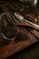 Delicious dried loose leaf tea is poured out of a glass jar on a wooden vintage brown table with a spoon, a cloth and a glass on a dark background photo