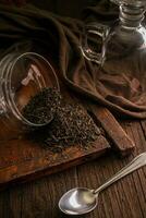 Dried leaf black tea is scattered from a glass jar on a wooden brown board with a spoon, a glass and a cloth on a dark background photo