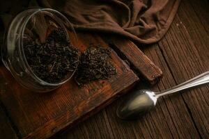 Green tea leaves in a glass jar with a spoon on a wooden brown board on a dark background. tea brewing time photo