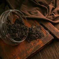 Delicious dried green tea leaves in a glass jar on a wooden brown board on a dark background, closeup photo