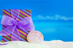 Striped gift box with bow and conch on sandy beach, behind sea. Vacations, relaxation, travel to hot countries. Christmas, New Year, birthday, Mother's and Women's Day, holiday. Copy space photo