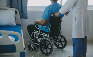 Rehabilitation of disabled people concept. Young physiotherapist helping senior male patient in wheelchair exercise at home. Handicapped elderly man training with dumbbells photo