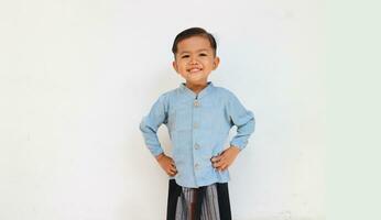 un linda y hermoso pequeño chico sonriente mientras levantamiento su mano en cintura. un chico vistiendo azul camisa y pareo de malasia foto