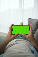 Young man at Home Uses Green Mockup Screen Smartphone in His Cozy Living Room.Vertical shot.Streaming service,watch videos online photo