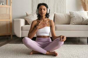 joven ajuste sano africano americano mujer en ropa de deporte sentado a hogar haciendo yoga, meditando y respiración en el Mañana. mental salud concepto foto