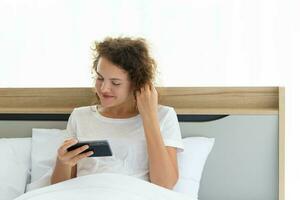 Young beautiful happy woman relaxing and using smartphone while lying in bed in the morning after waking up. photo