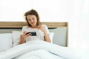Young beautiful happy woman relaxing and using smartphone while lying in bed in the morning after waking up. photo