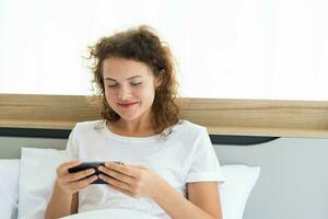 Young beautiful happy woman relaxing and using smartphone while lying in bed in the morning after waking up. photo