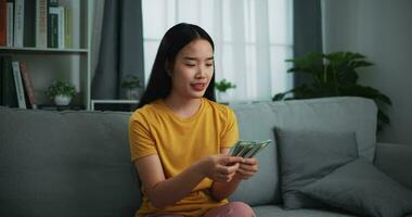 beeldmateriaal van jong Aziatisch vrouw tonen dollar Bij camera en glimlachen Aan sofa in de leven kamer Bij huis. video