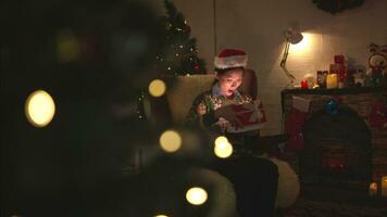 asiatico donna seduta su il sedia nel davanti di il Natale albero e apertura un' regalo scatola, allegro Natale e contento vacanze video