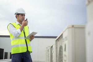 Asian maintenance engineer works on the roof of factory. contractor inspect compressor system and plans installation of air condition systems in construction. technology, online, mobile application. photo