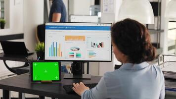 Businesswoman works at desk with greenscreen presented on modern tablet, looking at isolated copyspace template while she analyzes revenue data on pc. Worker uses chromakey display. Handheld shot. video