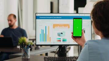 Adult holding phone with greenscreen in coworking space at office. Businesswoman working with smartphone showing isolated mockup template, analyzing chromakey layout on gadget. Tripod shot. video