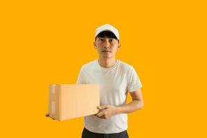 Young Asian delivery man wearing cap and white blank t-shirt holding parcel post box isolated on yellow background. express delivery service concept. photo