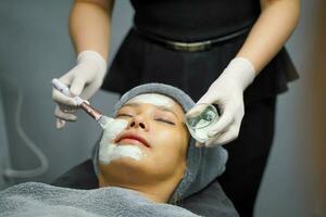 Closeup cosmetologist is applying facial white mask cream on woman client face in beauty clinic. photo