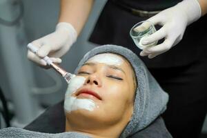 Closeup cosmetologist use cosmetic brush applying facial jelly's gel on woman client face in beauty clinic. photo