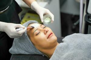 Closeup cosmetologist use cosmetic brush applying facial jelly's gel on woman client face in beauty clinic. photo