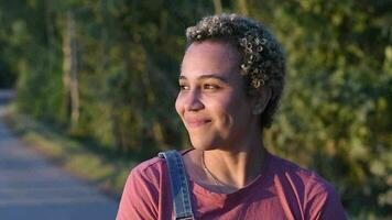 portrait de une Jeune brésilien millénaire fille avec court frisé cheveux des stands dans le forêt en riant gaiement et à la recherche à le coucher de soleil, haute qualité Full HD métrage video