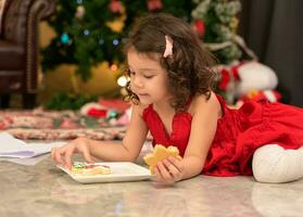 linda pequeño niños en pequeño niña en rojo vestir comiendo delicioso galletas a hogar. Navidad celebracion concepto foto