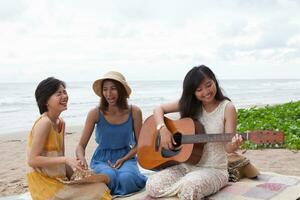 portrait group of young asian woman playing guitar in sea beach picnic party at sea side with happiness face emotion photo
