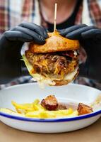 el concepto de rápido comida y para llevar alimento. un hombre en negro látex guantes sostiene un jugoso hamburguesa en su manos, mentiras cerca francés papas fritas en un metal plato a lo largo con queso salsa. de cerca. foto