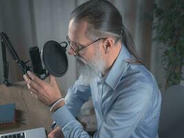 un masculino presentador con un barba prepara a transmitir su audio podcast utilizando un micrófono y ordenador portátil en su hogar estudio. de cerca foto