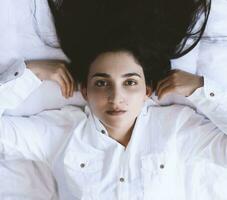 Beautiful woman lying in bed. Pretty woman with long brunette hairs lying in bed.High-angle view of a young happy girl dressed in a white shirt lies on a bed at home photo