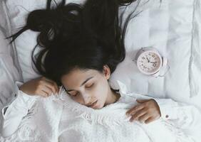 Beautiful black-haired woman sleeping in cozy bed. The alarm clock set at 7 a.m. morning. Female is going to wake up at 7 o'clock early morning. Top view photo