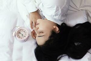 Beautiful black-haired woman sleeping in cozy bed. The alarm clock set at 7 a.m. morning. Female is going to wake up at 7 o'clock early morning. Top view photo
