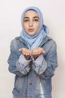 Portrait of stylish Muslim girl in jean jacket sending air kiss. Modern and young middle-eastern girl in hijab sending blowing air kiss. Lovely Persian woman expressing love photo