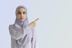 Cute Muslim woman with cheerful face expression points away with her forefinger, showing to copy space on the corner. Portrait of Muslim prayer woman in hijab photo