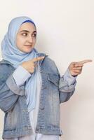 Cute Muslim woman with cheerful face expression points away with her forefinger, showing to copy space on the corner. Portrait of Muslim prayer woman in hijab photo