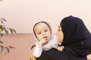 joven musulmán mujer en hijab participación su 1 año bebé chico, abrazando y besando contento musulmán madre en abaya ropa Beso y jugar con su pequeño niño foto