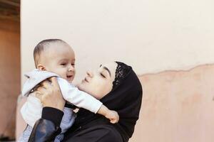 joven musulmán mujer en hijab participación su 1 año bebé chico, abrazando y besando contento musulmán madre en abaya ropa Beso y jugar con su pequeño niño foto