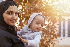 joven musulmán mujer en hijab participación su 1 año bebé chico, abrazando y besando contento musulmán madre en abaya ropa Beso y jugar con su pequeño niño foto