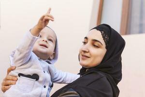 Little toddler kid in hands of her mother, Happy Muslim family. Baby boy pointing with his finger to upwards showing something to her mother photo