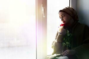 Young Muslim woman sits on the window sill with flower bouquet in hands. Depressed middle-eastern female near the window at home. Lonely cheerless girl feeling stressed and waiting photo