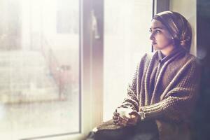 Young Muslim woman sits on window sill and looking away. Depressed middle-eastern female near the window at home. Lonely cheerless girl feeling stressed and waiting photo