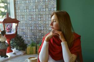 A girl with blond hair in a red scarf sits at a window decorated for Christmas. Portrait of a young woman on Christmas Eve, waiting for the holiday. photo