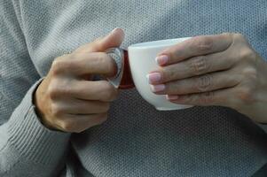 A woman in a gray sweater holds a white cup of tea or coffee. Close-up of female hands with a cup. photo