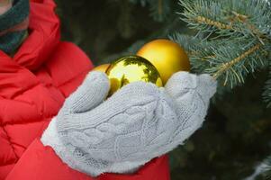 Female hands in warm knitted gray mittens hold golden Christmas balls on the background of a Christmas tree. Preparation for the holiday. photo